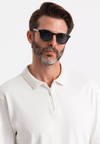 Schwarze Sonnenbrille mit runden Gläsern und einer markanten grauen Brücke, getragen zu einem schlichten weißen Polo-Shirt mit Reißverschluss. Textilstruktur und klassisches Design.
