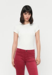 Femeie cu păr închis la culoare, de lungime medie, purtând un top alb cu mâneci scurte și pantaloni roșii cu talie înaltă, stând pe un fundal alb.