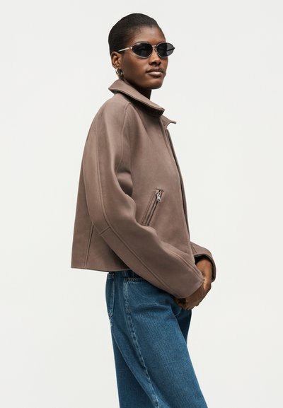 Γυναίκα με στρογγυλά μαύρα γυαλιά ηλίου, χρυσούς κρίκους σκουλαρίκια, μια ζακέτα σε χρώμα ταουπ με φερμουάρ και μπλε τζιν, ποζάρει σε ανοιχτό φόντο.