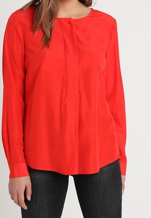 Femme portant un chemisier rouge vif à manches longues avec un col boutonné, associé à un pantalon noir, debout devant un fond uni.