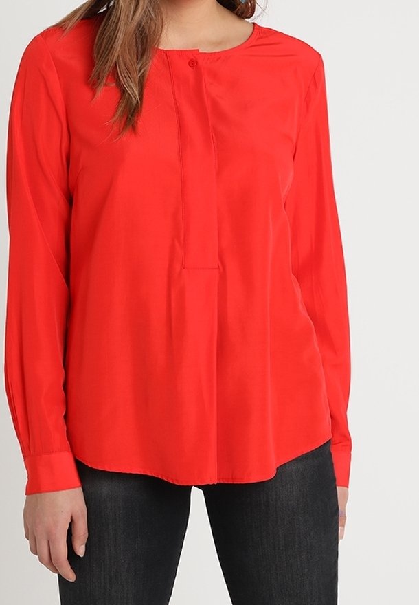 Femme portant un chemisier rouge vif à manches longues avec un col boutonné, associé à un pantalon noir, debout devant un fond uni.