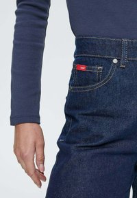 Personne portant un jean bleu foncé avec une étiquette rouge visible sur la poche et un haut bleu marine à manches longues, la main détendue à ses côtés.