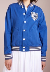 Osoba v modré zapínací bundě s pruhovanými manžetami a lemem, která má našitou nášivku lva, v kombinaci s bílou skládací sukní.