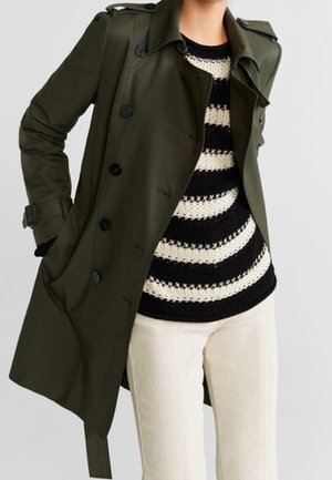 Personne portant un trench-coat vert foncé par-dessus un pull rayé noir et blanc et un pantalon clair, debout avec une main dans la poche du manteau.