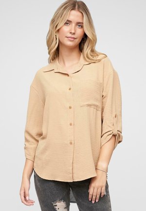 Camisa beige de botones en un tejido texturizado, con cuello, mangas largas y un bolsillo en el pecho. Combinada con jeans oscuros desgastados.