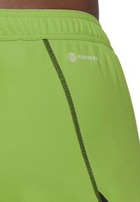Grüne Sportshorts aus glattem synthetischem Material, mit schwarzen Nähten, einer Außentasche und einem Logo auf der Rückseite.