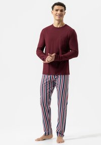 Rood shirt met lange mouwen en een zak, gecombineerd met blauw, wit en rood verticaal gestreepte pyjamabroek. Model staat blootsvoets en glimlacht.