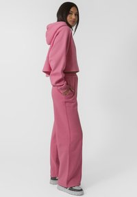 Un hoodie court rose avec des manches longues et une capuche, associé à un pantalon de survêtement large assorti. Le matériau semble doux et texturé.