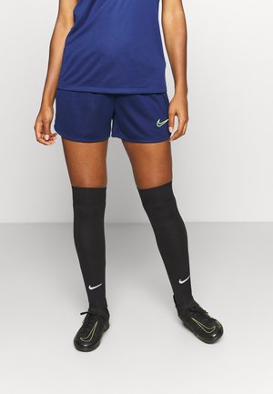 Person mit marineblauen Sportshorts und Oberteil, schwarzen knielangen Socken und schwarzen Nike Fußballschuhen, steht vor einem schlichten Hintergrund.