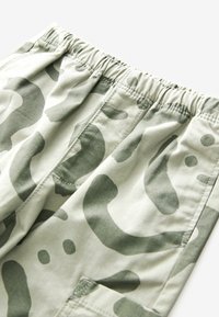 Hellgrüne Camouflage-Hosen mit elastischem Bund, dekoriert mit abstrakten dunkelgrünen Formen, sichtbaren Nähten und Seitentaschen.