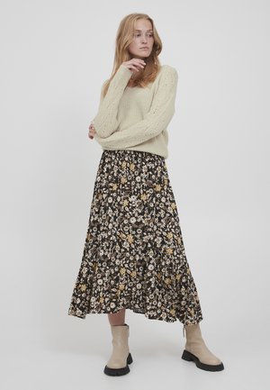 Strickpullover in Creme mit strukturiertem Muster, kombiniert mit einem schwarzen Rock mit Blumenmuster, der gelbe und weiße Blumen zeigt. Beigefarbene Stiefeletten runden das Outfit ab.