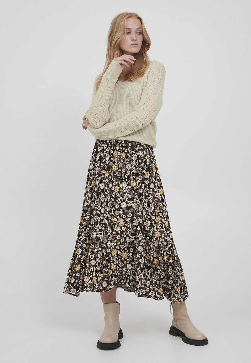 Strikket cremefarvet sweater med et tekstureret mønster, sat sammen med en sort blomsterkjol med gule og hvide blomster. Beige ankelstøvler fuldender looket.