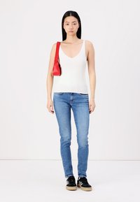 Top in cotone bianco a costine, jeans attillati blu, sneakers nere con suola beige, piccola borsa rossa. Sfondo semplice, modello in piedi rivolto in avanti.