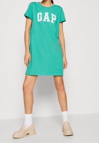 Grünes Kurzarmkleid mit einem weißen "GAP"-Logo, lockerer Passform und weichem Material. Kombiniert mit klobigen beigefarbenen Plattformschuhen und weißen Socken.