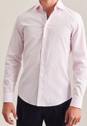 Mann trägt ein hellrosa langärmeliges Hemd mit Knöpfen und kontrastierendem innerem Kragen, kombiniert mit dunkler Hose.