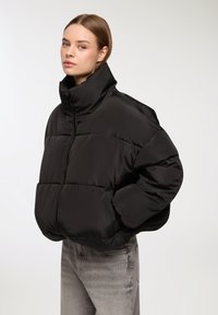 Giacca a vento nera con collo alto, presenta sezioni trapuntate, zip frontale e vestibilità oversize, abbinata a jeans grigi chiari.