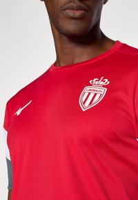 Camiseta atlética roja con cuello redondo, que presenta un diseño de logo blanco y corona. El material parece suave, tiene mangas cortas y acentos de color.