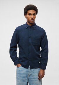 Texturált anyagból készült sötétkék gombos ing, klasszikus gallérral, elülső zsebbel és egyenes szabással, világoskék farmerrel párosítva.