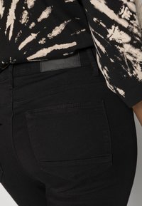 Jeans de ganga preta com um design simples, bolsos traseiros sem ferragens visíveis e uma textura lisa. Acompanhados por uma blusa com padrão.