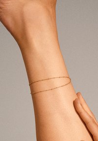Un bracelet double chaîne en or repose sur un poignet, présentant un design délicat et fin avec une finition lisse et un éclat subtil contre la peau.