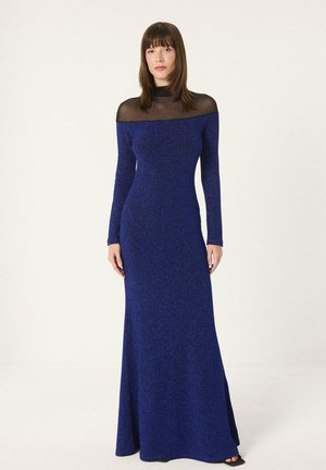 Kobieta w niebieskiej, długiej do ziemi sukience z długimi rękawami i prześwitującym czarnym wysokim kołnierzem, stojąca na jednolitym jasnym tle.