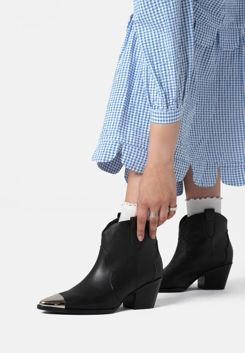 Botas de tobillo de cuero negro con puntas afiladas, que cuentan con tapas metálicas plateadas y lados texturizados, usadas con calcetas blancas con relieve y un vestido de cuadros azul.