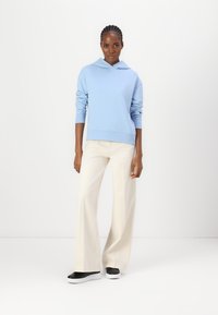 Felpa in cotone azzurro chiaro con tasca frontale, abbinata a pantaloni larghi color crema e sneakers bianco e nere. Il modello si trova di fronte a uno sfondo semplice.