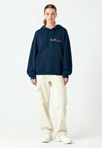 Felpa oversize blu navy con tasca frontale e testo ricamato rosso, abbinata a pantaloni cargo beige chiaro e sneakers bianche.