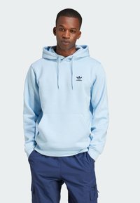 Lichtblauwe hoodie met een kangoeroezak, verstelbare trekkoordcapuchon en een marineblauw Adidas-logo op de borst. Gecombineerd met marineblauwe cargobroek.