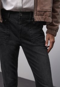 Černé džíny z denimu s úzkým střihem, s kapsami na zip a strukturovaným pasem. Textura působí hladce a odolně.