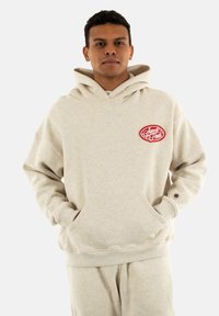 Sudadera oversized de color beige claro con un bolsillo frontal y un logotipo bordado en rojo. Tela suave, mangas largas y puños acanalados.