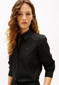 Ung kvinne med krøllete hår, iført en svart skjorte med knapper og en liten logo på brystet, står foran en hvit bakgrunn.