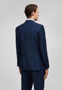 Veste de costume bleu marine avec une coupe ajustée. Présente un tissu lisse, une fente simple au dos et trois boutons à chaque poignet.