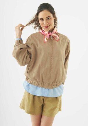 Young woman wearing beige jacket, light blue shirt, mustard shorts, and pink patterned neck scarf, holding a curl of her hair.