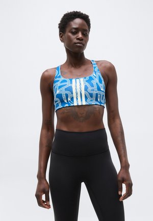 Γυναίκα που φοράει μπλε αθλητικό σουτιέν με λευκές ρίγες και μαύρο κολάν με ψηλή μέση, στέκεται μπροστά σε έναν απλό φόντο.