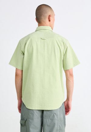 Man with shaved head wearing light green short-sleeve shirt and grey cargo pants, standing facing away against a white background.