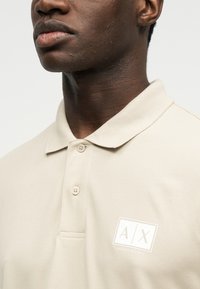 Κοντινό πλάνο ενός ατόμου που φοράει μπεζ polo μπλούζα Armani Exchange με γιακά και λογότυπο στο αριστερό μέρος του στήθους.