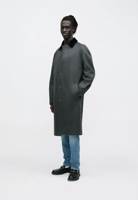 Grauer Mantel aus glattem Material mit schwarzem Kragen, Frontknöpfen und Seitentaschen, kombiniert mit blauer Jeans und schwarzen Schuhen.