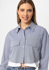 Lys blå chambray skjorte med knaplukning fortil, to brystlommer og en hvid krave, der ses nedenunder. Bæres med lyse denimbukser.
