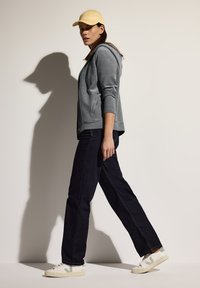 Felpa grigia con zip, jeans scuri, cappellino giallo e sneakers bianche con dettagli verdi. La felpa ha una texture liscia e un cappuccio.