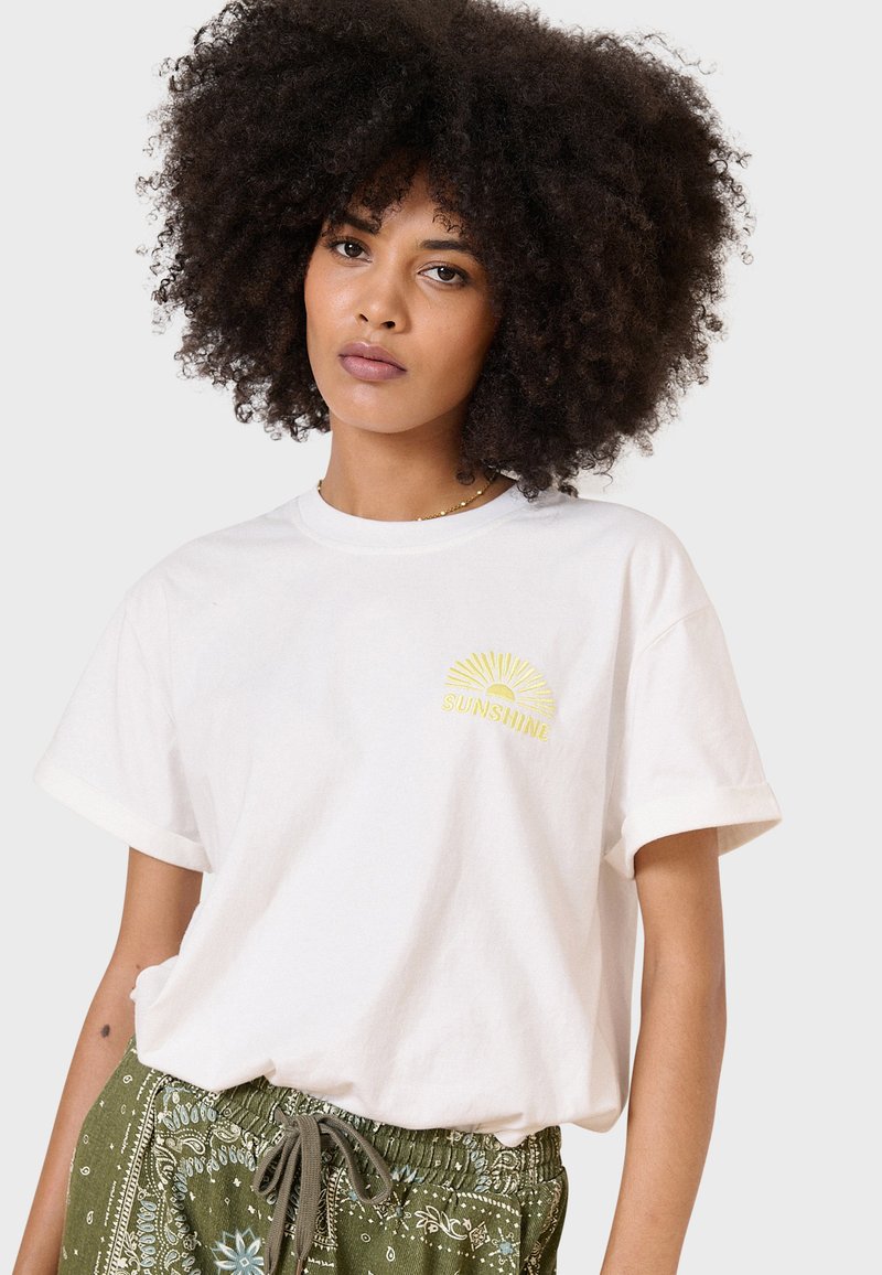 Weißes Baumwoll-T-Shirt mit umgeschlagenen Ärmeln, mit einem gelben Sonnenmotiv und dem Wort "SUNSHINE" auf der linken Brust gestickt.