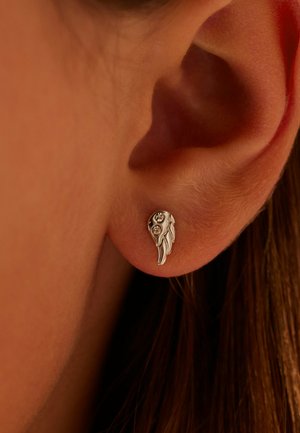Ohrringe aus Sterlingsilber in Flügelform mit zwei kleinen klaren Edelsteinen, strukturierten Details, die an einem Ohr einer Person mit braunem Haar befestigt sind.