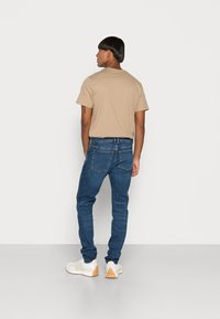 T-shirt bege e jeans azul escuro usados com ténis brancos. Os jeans apresentam um design clássico com bolsos traseiros e um ligeiro elástico.