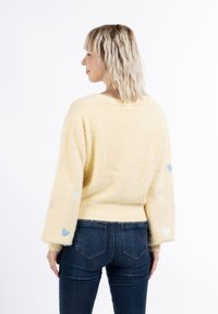 Maglione giallo sfocato con una vestibilità comoda, caratterizzato da toppe a forma di cuore colorate sulle maniche. Abbinato a jeans blu scuro, che mostrano un look informale.