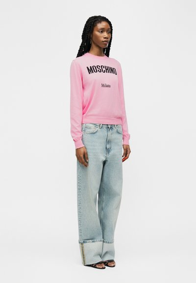 Femme portant un sweat-shirt rose Moschino Milano, un jean large bleu clair avec revers roulés, et des sandales noires à bouts ouverts, debout devant un fond blanc.