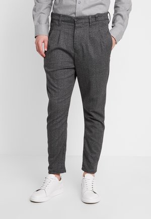 Mężczyzna w szarych, dopasowanych spodniach, jasnoszarej koszuli na guziki i białych trampkach, stojący z rękami w kieszeniach na jednolitym tle.