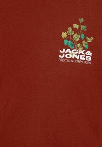 Sarkana apakšveļa ar apaļu kakla izgriezumu, izgatavota no mīksta kokvilnas, ar zaļu lapas un globusa motīvu, kā arī tekstu "JACK & JONES" un "IZVEIDOTS KOPENHĀGENĀ".