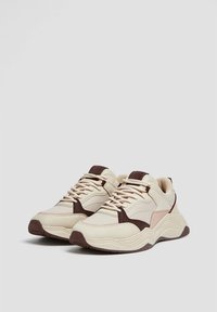 Scarpe sportive beige con accenti marroni scuri, pannelli testurizzati e un design della suola curvata. Presentano lacci e supporto per la caviglia imbottito.