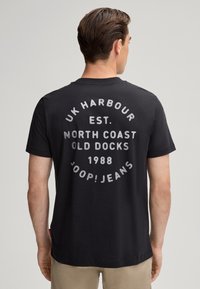 T-shirt en coton noir avec un imprimé graphique rond au dos comportant du texte en blanc : "UK HARBOUR EST. NORTH COAST OLD DOCKS 1988 JOOP! JEANS."
