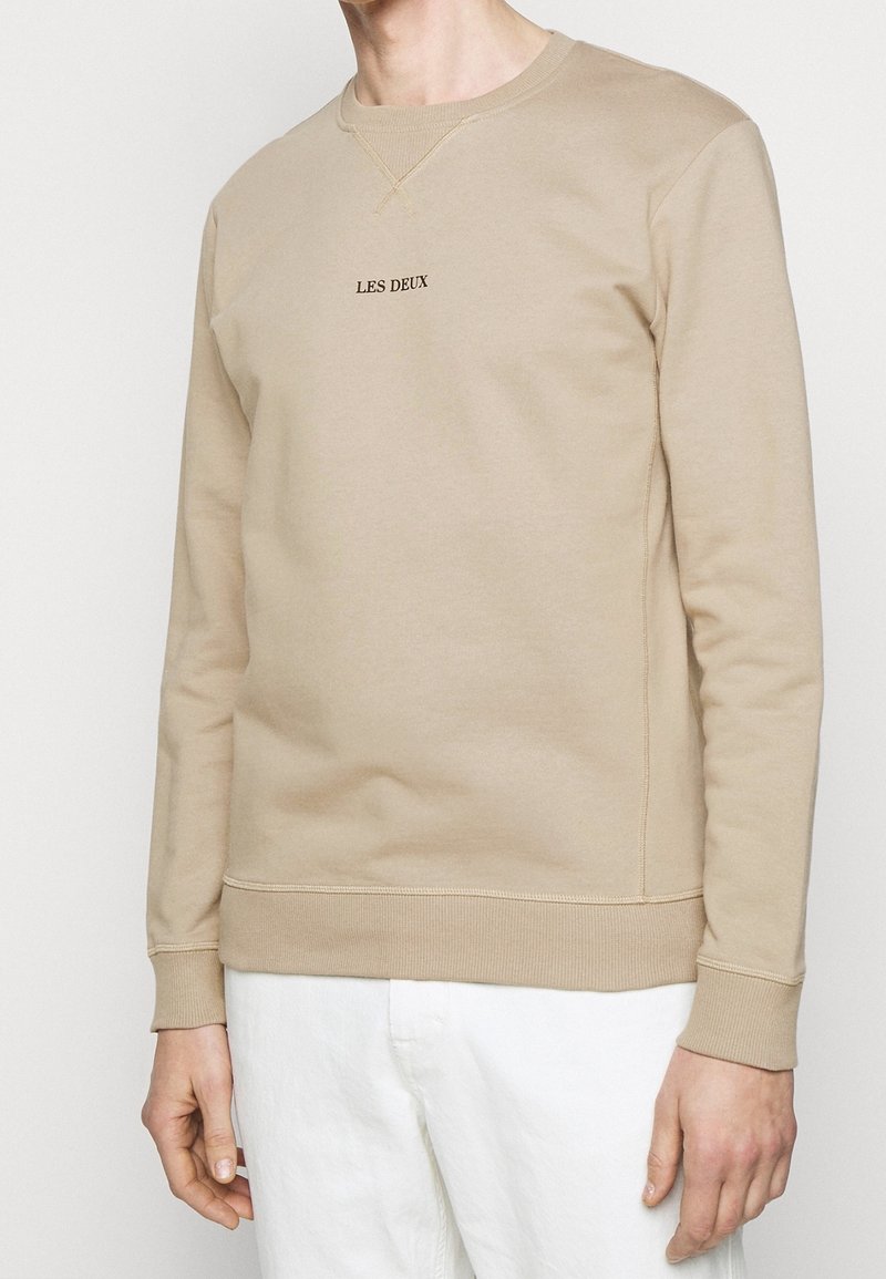 Sweat-shirt beige à manches longues avec le texte "LES DEUX" sur la poitrine, porté par une personne portant un pantalon blanc, cadré au niveau du cou et du milieu de la cuisse.